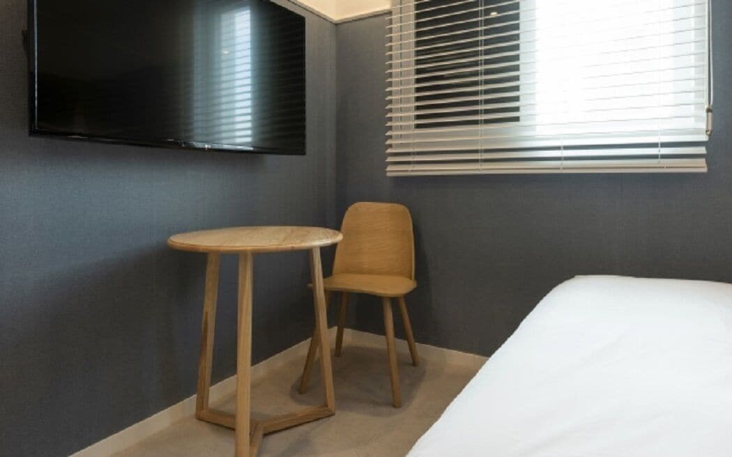 Een hotelkamer met een bed, een klein rond tafeltje, een stoel, een aan de muur bevestigde televisie en een raam met jaloezieën.