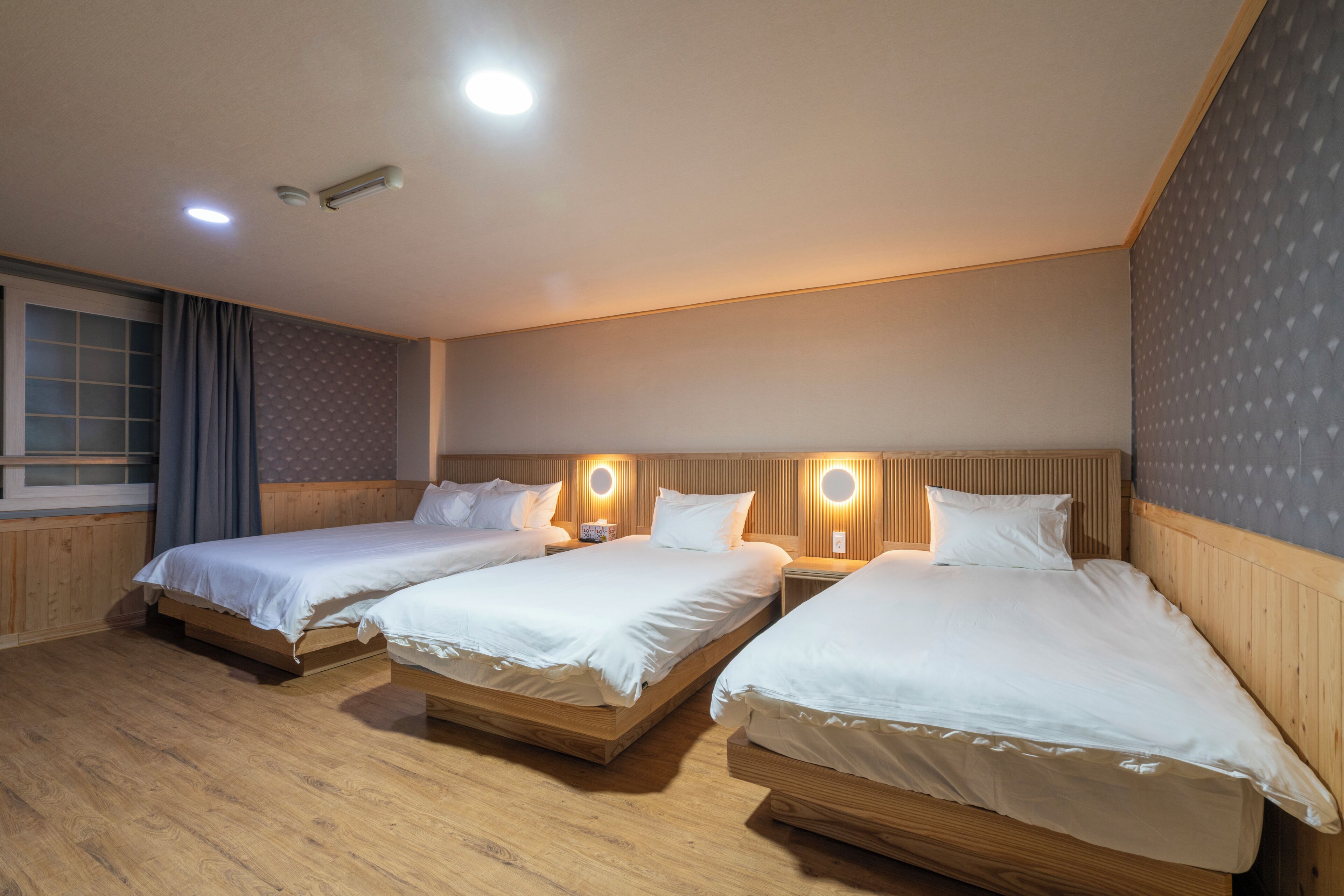 A hotel room with two single beds, wooden flooring, a window with curtains, and wall-mounted lighting.