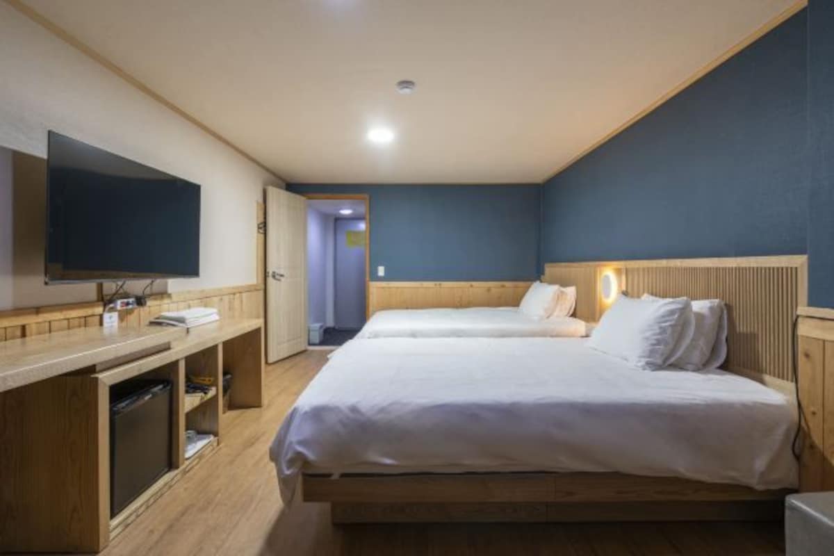 Une chambre d’hôtel avec un lit en bois, un téléviseur à écran plat, un bureau avec un ordinateur et une porte donnant sur une autre pièce.