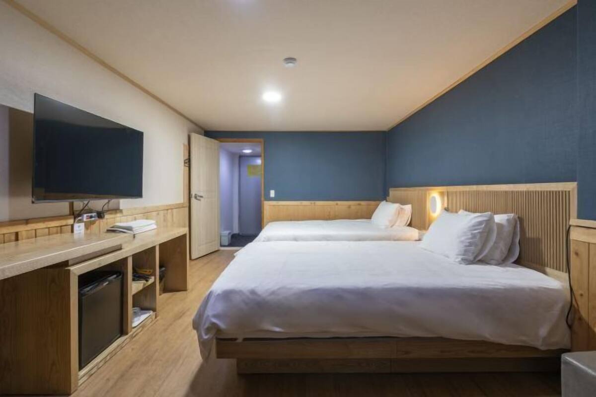 Ett hotellrum med en säng i trä, en platt-TV, ett skrivbord med en dator och en dörr som leder till ett annat rum.