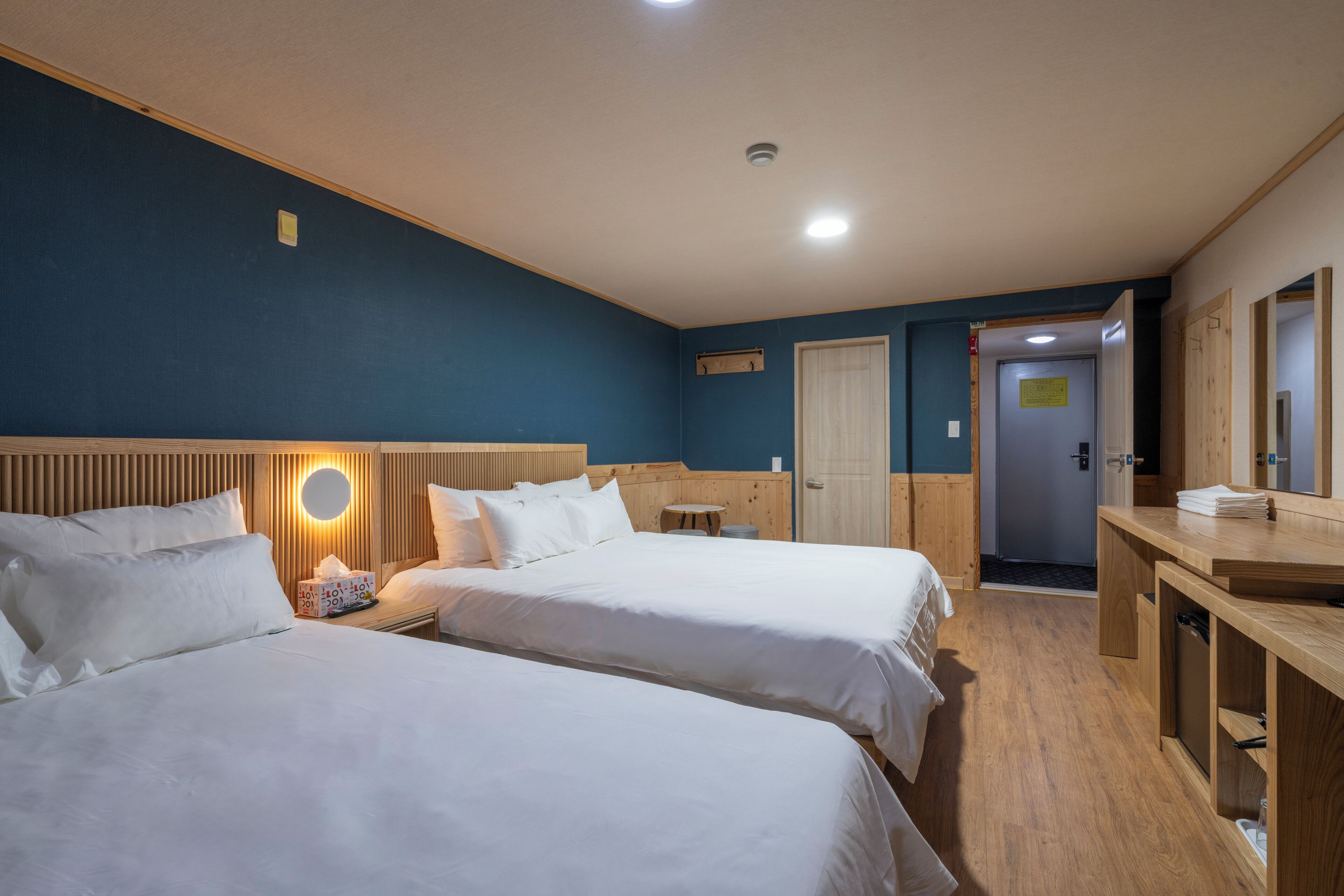 Habitación de hotel con dos camas, cabecero de madera, mesita de noche con lámpara, una pequeña mesa redonda, escritorio de madera y espejo.