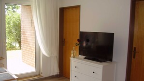 Ein Zimmer mit Fernseher auf einem weißen Schrank, einer Holztür und einem fenster mit Vorhängen.