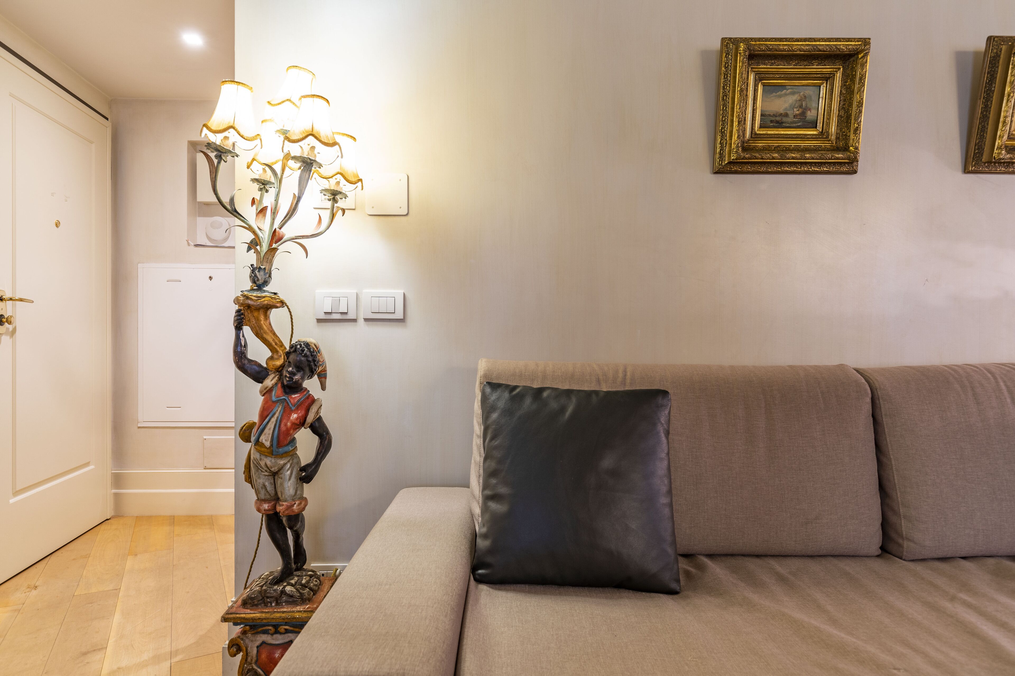 Un salón con un sofá color beige, un cojín marrón oscuro, un aplique decorativo en la pared y un cuadro enmarcado.