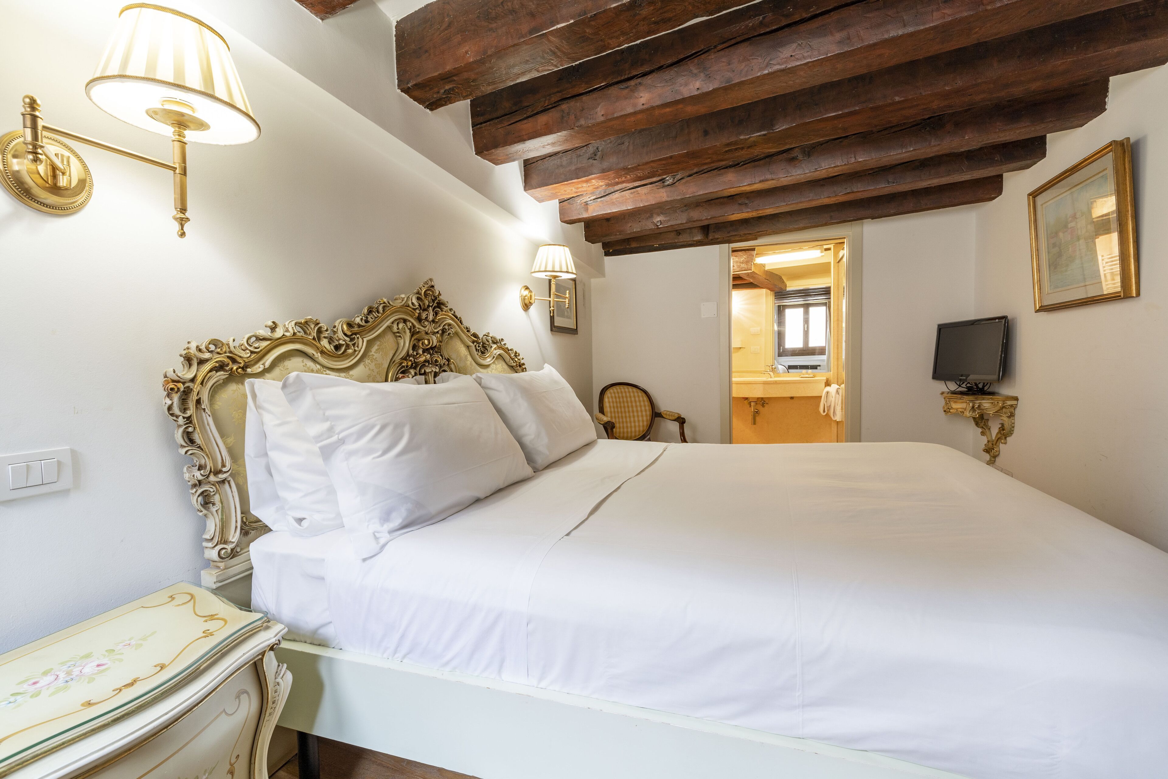 Ein Schlafzimmer mit einem großen Bett, einem verzierten Kopfteil, Wandlampen und einem Holzbalken an der Decke.