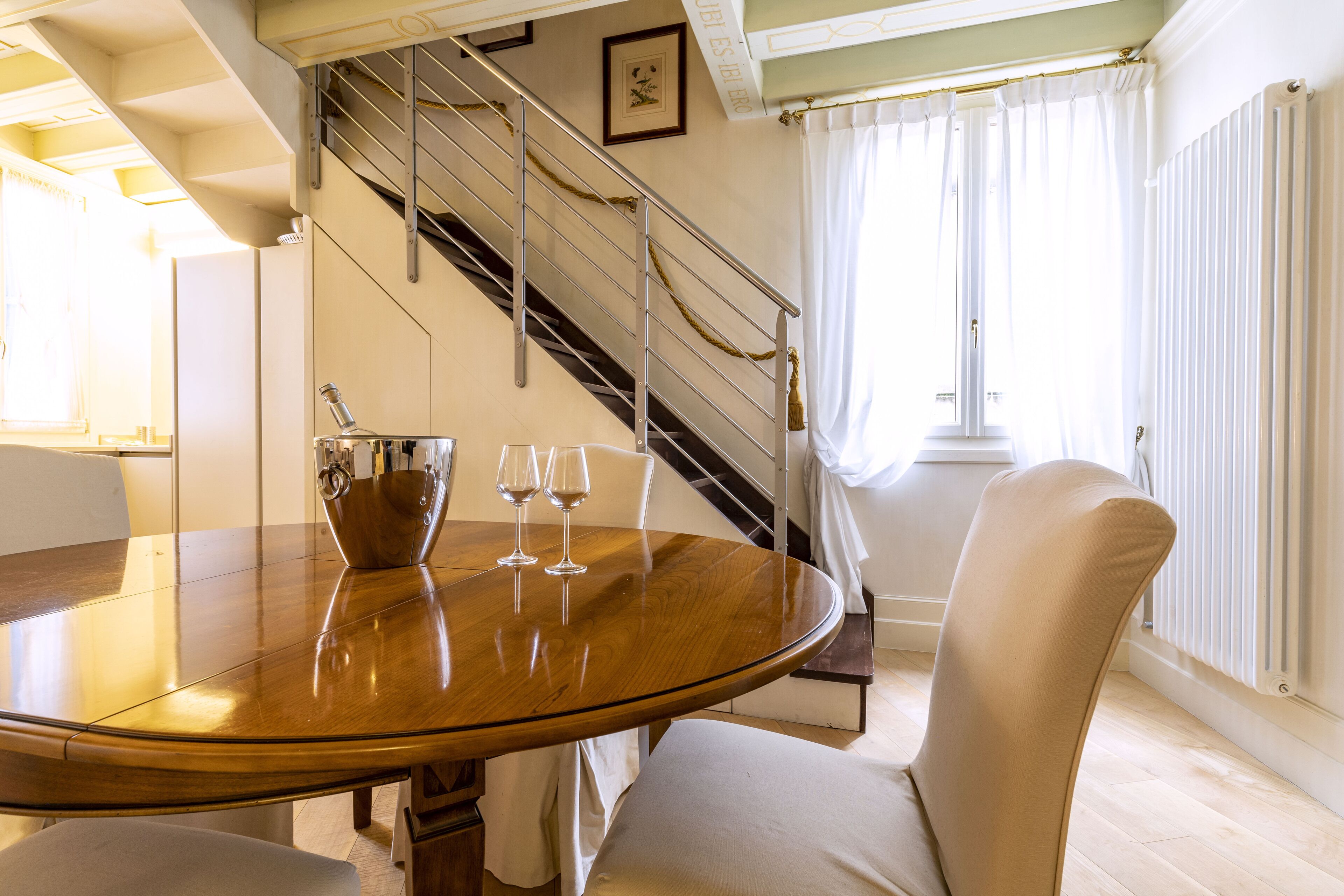 A dining area with a wooden table, two chairs, and a vase with wine glasses. A staircase with a metal railing is visible in the background.