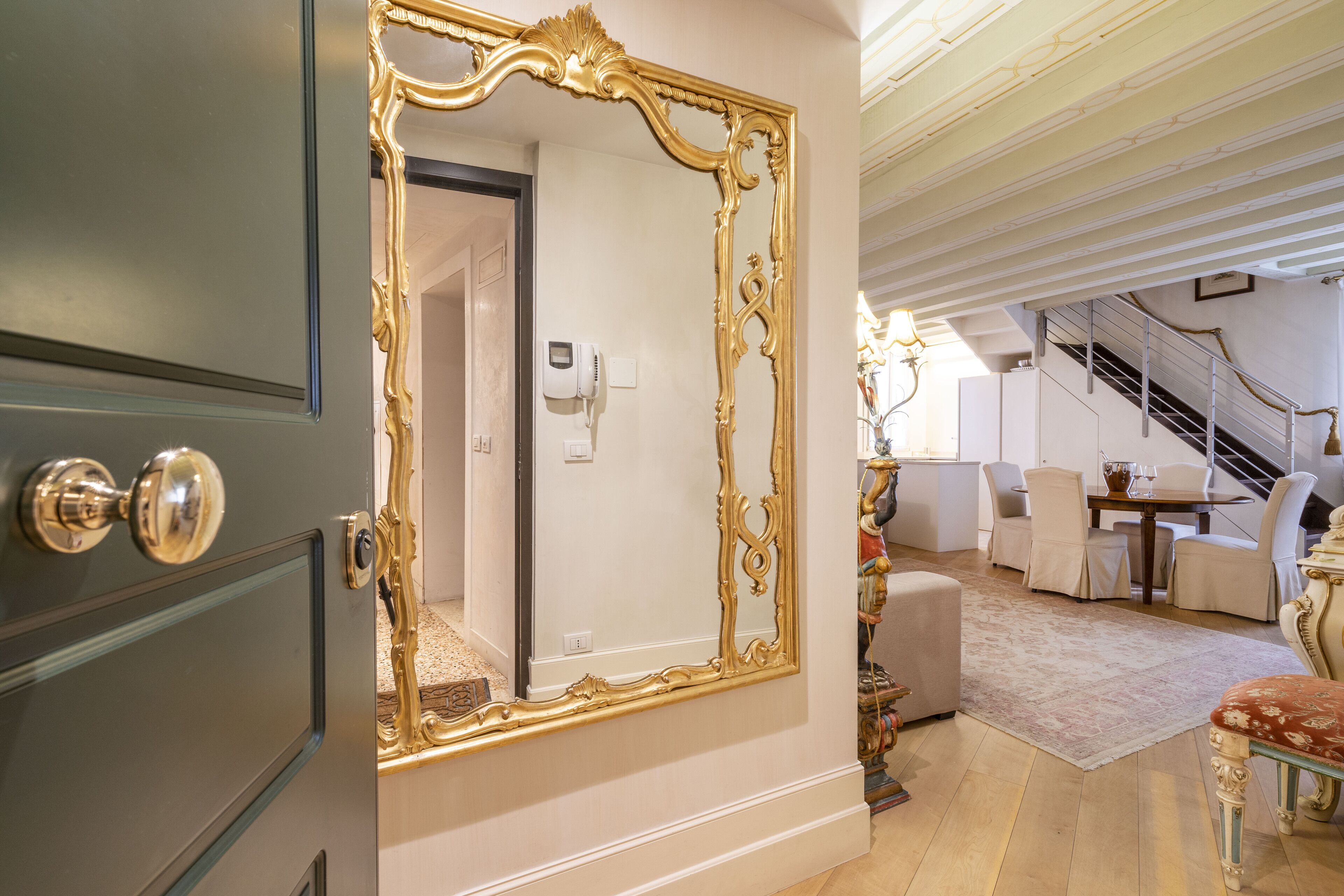 Una habitación con un espejo grande y ornamentado, una escalera y un comedor con mobiliario blanco.