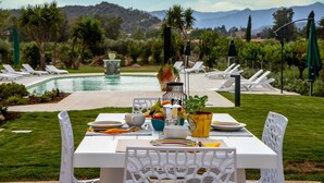 Une table à manger dressée pour quatre personnes, avec des chaises blanches, une piscine et une chaîne de montagnes en arrière-plan.
