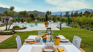 Une table à manger installée à l’extérieur, avec une piscine et des montagnes en arrière-plan.
