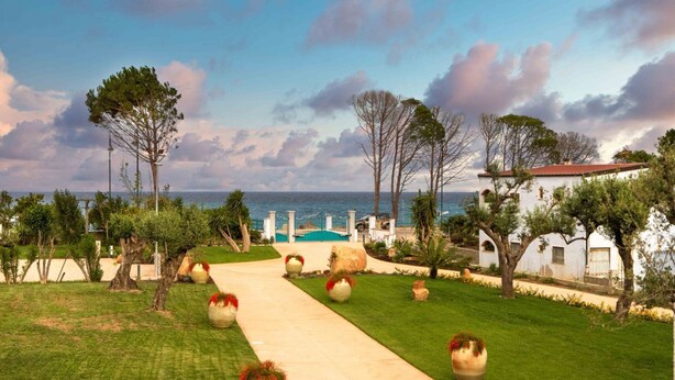 Un chemin mène à un bâtiment blanc surmonté d’un toit rouge, entouré de verdure et d’arbres, avec une vue sur la mer en arrière-plan.