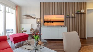A modern living room with a glass coffee table, red sofa, and a flat-screen TV mounted on a wooden panel wall. - FW 18, apartments on the beach - max. 4 persons - FeWo Am Strand / (Kühlungsborn)