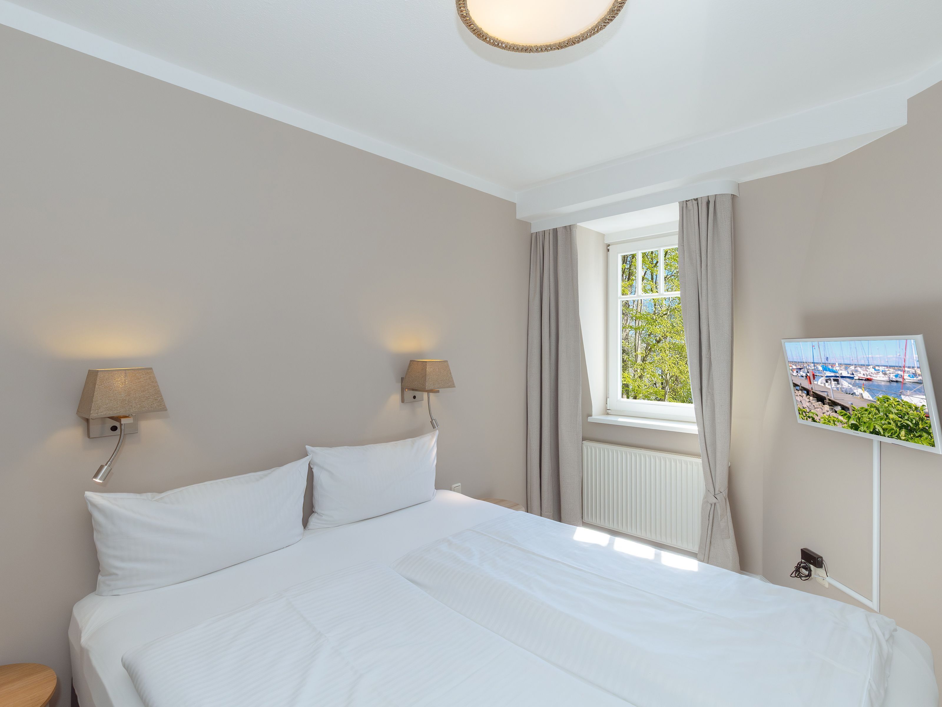 A hotel room with a bed, two wall-mounted lamps, a window with curtains, and a TV on the wall.