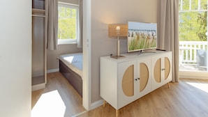Ein Schlafzimmer mit einem Bett, einem Fernseher auf einer weißen Konsole, einem Fenster mit Vorhängen und Blick auf Bäume.