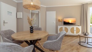 Ein modernes Wohnzimmer mit einem runden Holztisch, grau gepolsterten Sesseln, einer weißen Kommode mit runden Ausschnitten, einem Fernseher, der einen Sonnenuntergang zeigt, und einer hängenden Lampenschirm aus Gewebe.