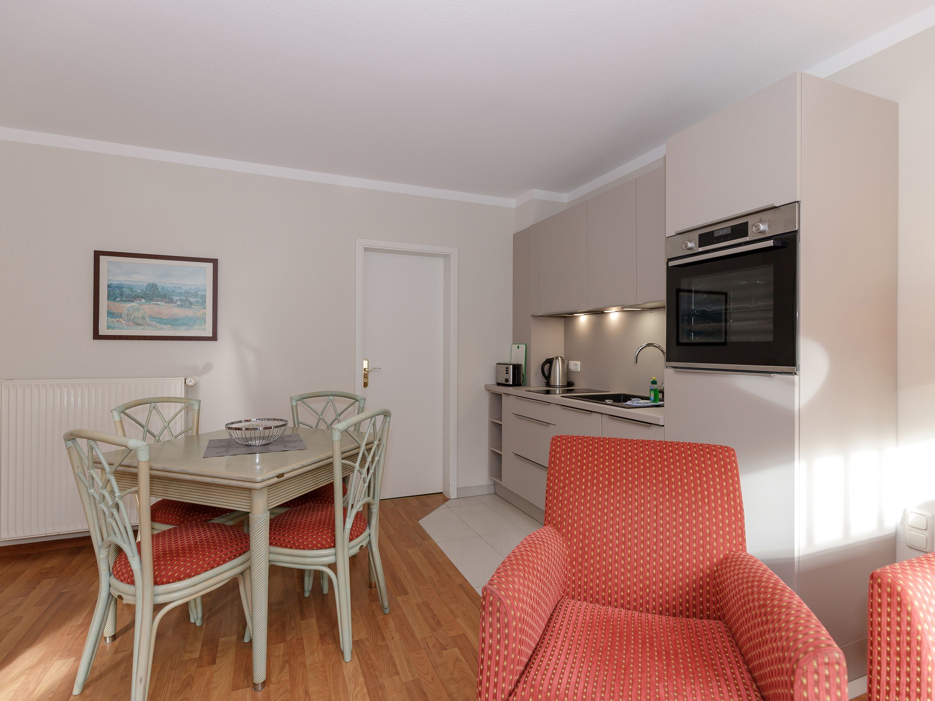 A compact kitchen with a dining area, featuring a table, chairs, a red patterned sofa, and a framed landscape painting on the wall.