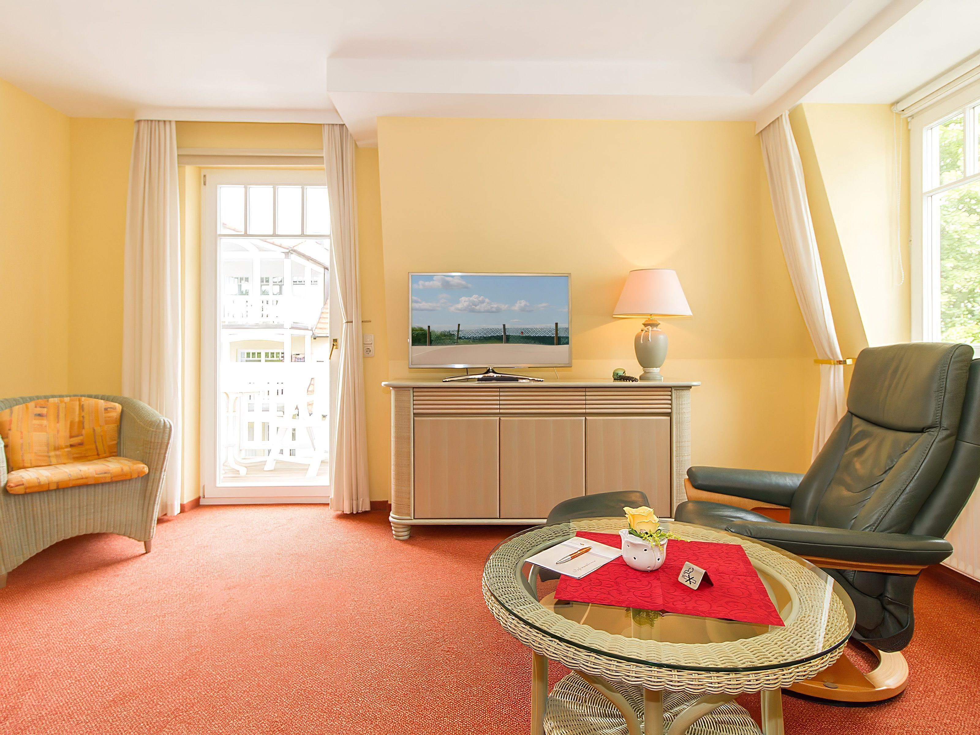 Une chambre d’hôtel équipée d’une télévision, d’une lampe, d’une table en verre avec un set de table rouge, d’une chaise et d’un canapé.