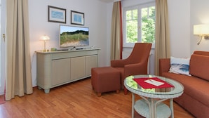 Een kamer met een televisie, een bank, een stoel, een klein tafeltje en een raam met gordijnen.