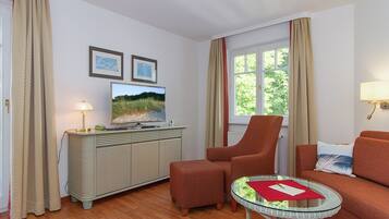 Une pièce comprenant une télévision, un canapé, un fauteuil, une petite table et une fenêtre avec des rideaux.