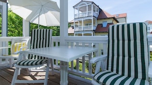 Een overdekt terras met witte meubels en gestreepte kussens, met uitzicht op een woongebouw met een balkon.