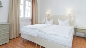 Een tweepersoonsbed met twee kussens, een nachtkastje, een dressoir en een raam met gordijnen.