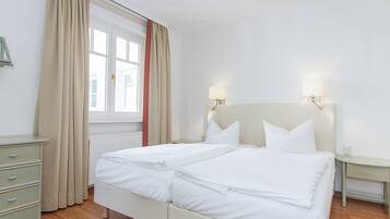 A double bed with two pillows, a bedside table, a nightstand, a lamp, a dresser, and a window with curtains.