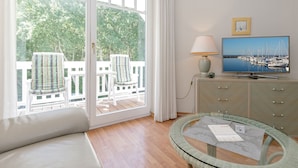 Una habitación con sofá, mesa de vidrio, televisión y vista a un balcón con sillas.