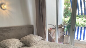 Een netjes opgemaakt bed met beddengoed met een patroon en een opgevouwen handdoek, een wandlamp en uitzicht op een buitenbalkon met een gestreept zonnescherm.