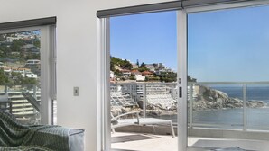A balcony with a view of a coastal town, a white chair, and a grey cushioned bench. - Seacliffe 502 (Cape Town)