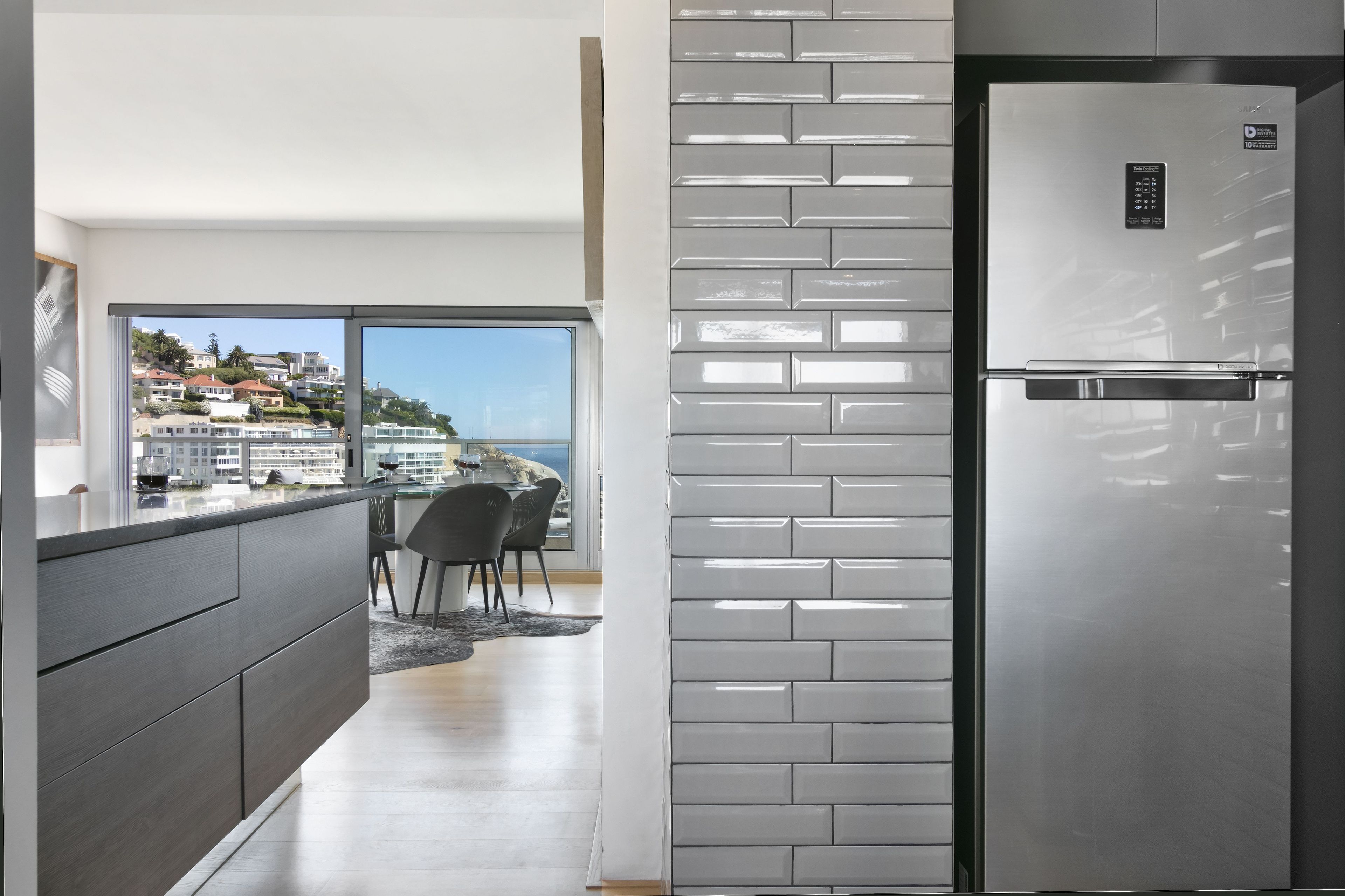 Uma cozinha moderna com frigorífico de aço inoxidável, uma janela ampla com vista para uma cidade costeira e um espaço de refeições com vista.