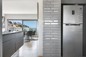 Una cocina moderna con un refrigerador de acero inoxidable, un ventanal con vistas a una ciudad costera y un comedor con panorama.