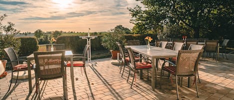 An outdoor patio with a table and chairs, surrounded by greenery and a clear sky.