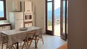 Une cuisine moderne avec des meubles blancs, une table à manger avec des chaises, et une vue sur la mer par la porte ouverte.