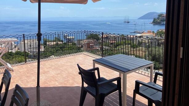 Un balcon avec une table et des chaises, un parasol beige, et une vue sur la mer et les bâtiments éloignés.