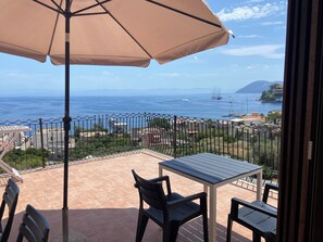 A balcony with a table and chairs, a beige umbrella, and a view of the sea and distant buildings. - House (Lipari)