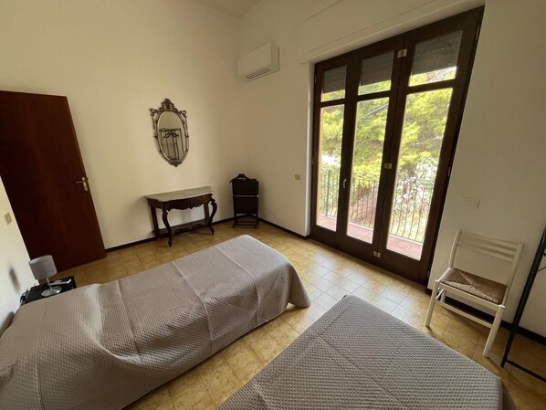 A room with two beds, a wooden door, a bench, a table, and a large window with a view of greenery.
