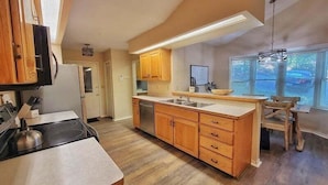 A kitchen with wooden cabinets, a central island, a dining area with a table and chairs, and a large window providing natural light. - Fairway Mountain Retreat-Views-5min to Downtown (Waynesville)