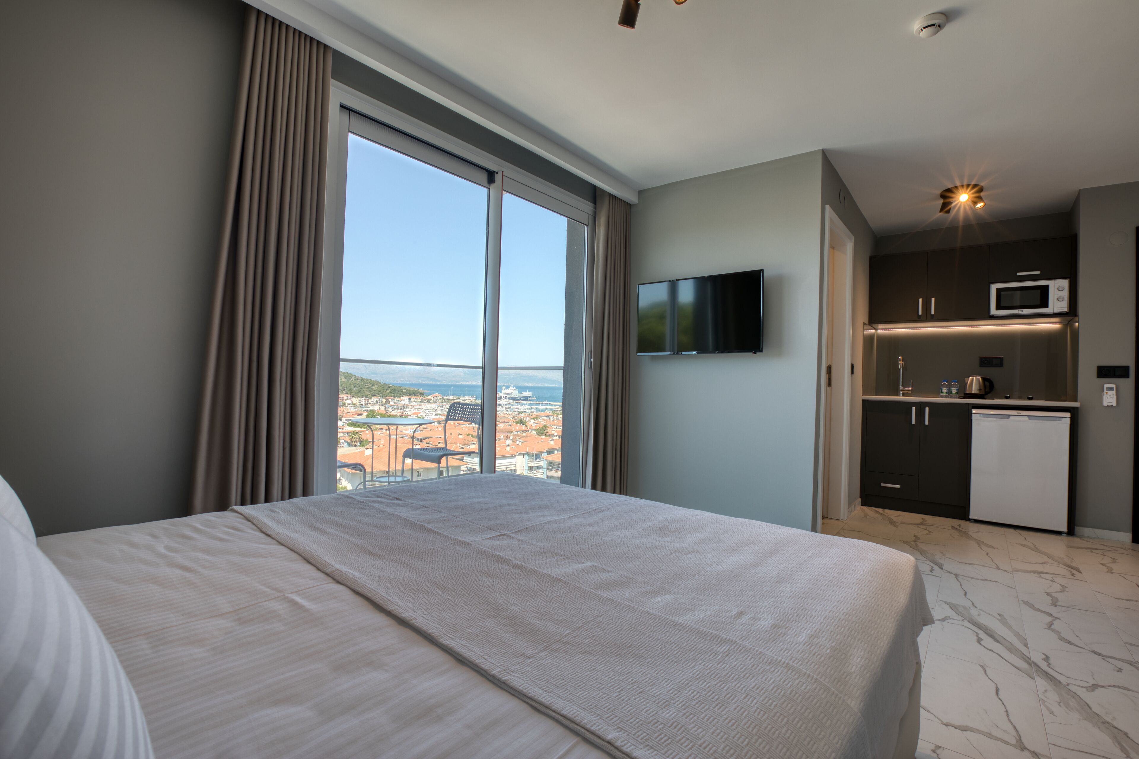 A hotel room with a large bed, a TV mounted on the wall, a kitchenette, and a view of the city through the window.