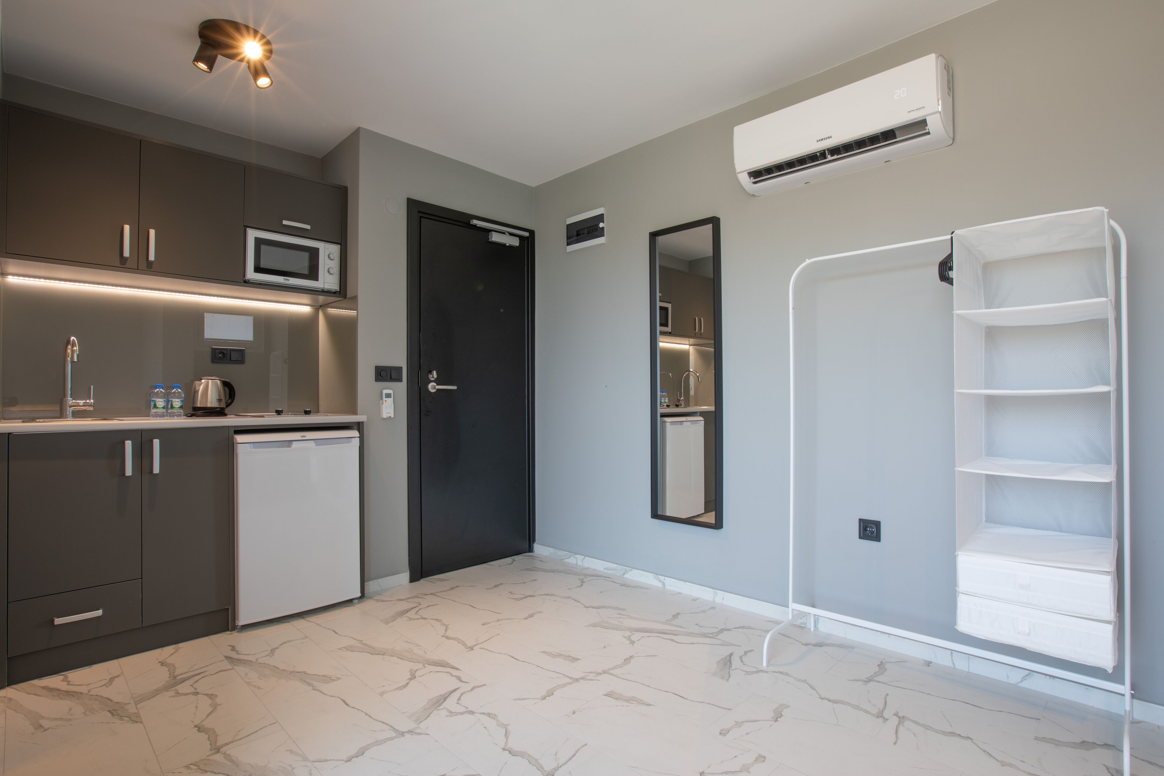 A modern kitchen with grey cabinets, a microwave, and a dishwasher. There is a black door, a white shelf unit, and a marble floor.