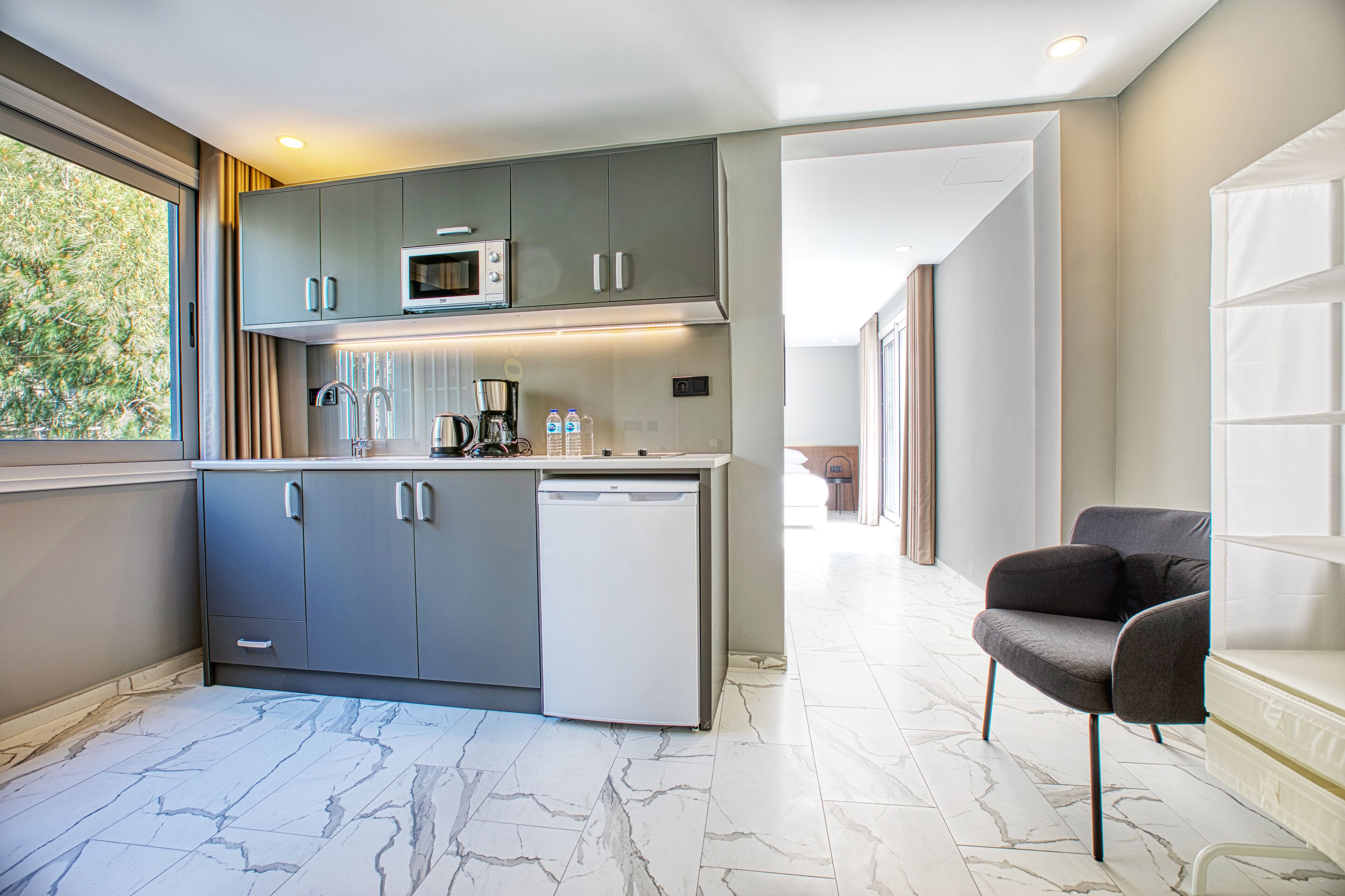 A modern kitchen with grey cabinets, a microwave, and a dishwasher. There is a marble floor and a grey armchair in the adjacent living area.