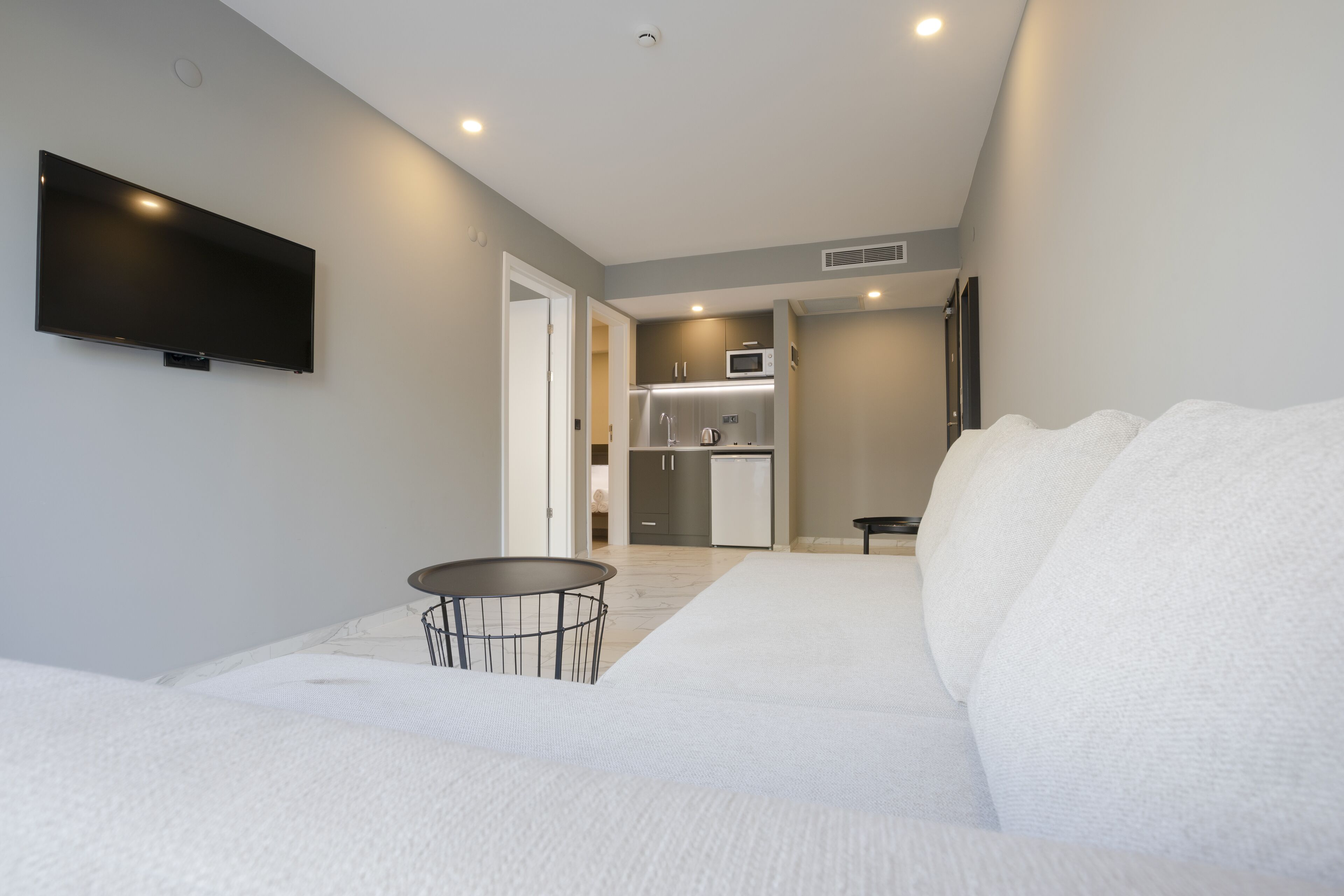 A modern hotel room with a large bed, a flat-screen TV mounted on the wall, a small round table, and a kitchenette in the background.
