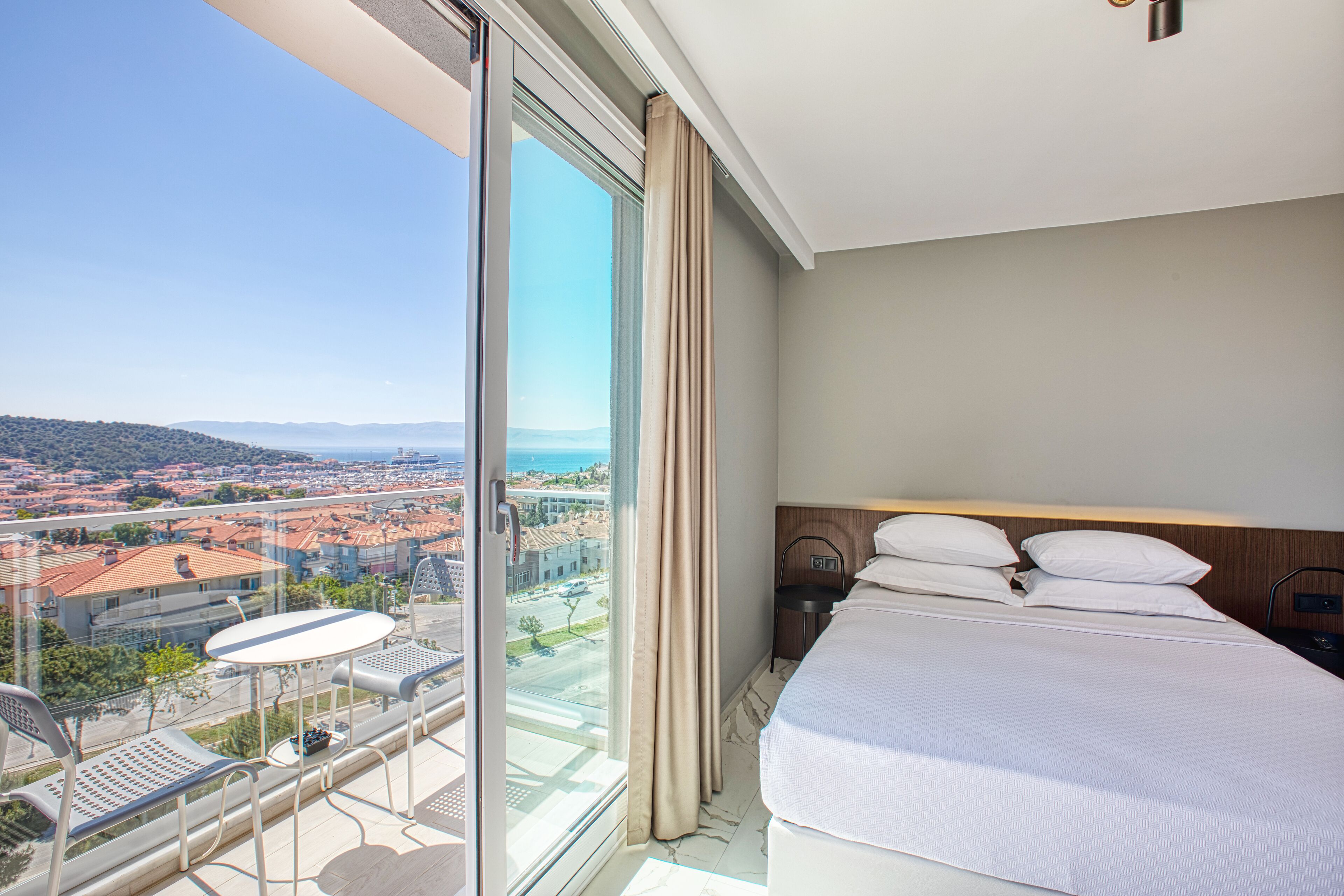 A hotel room with a large sliding glass door leading to a balcony, a bed with white linens, and a view of a cityscape.