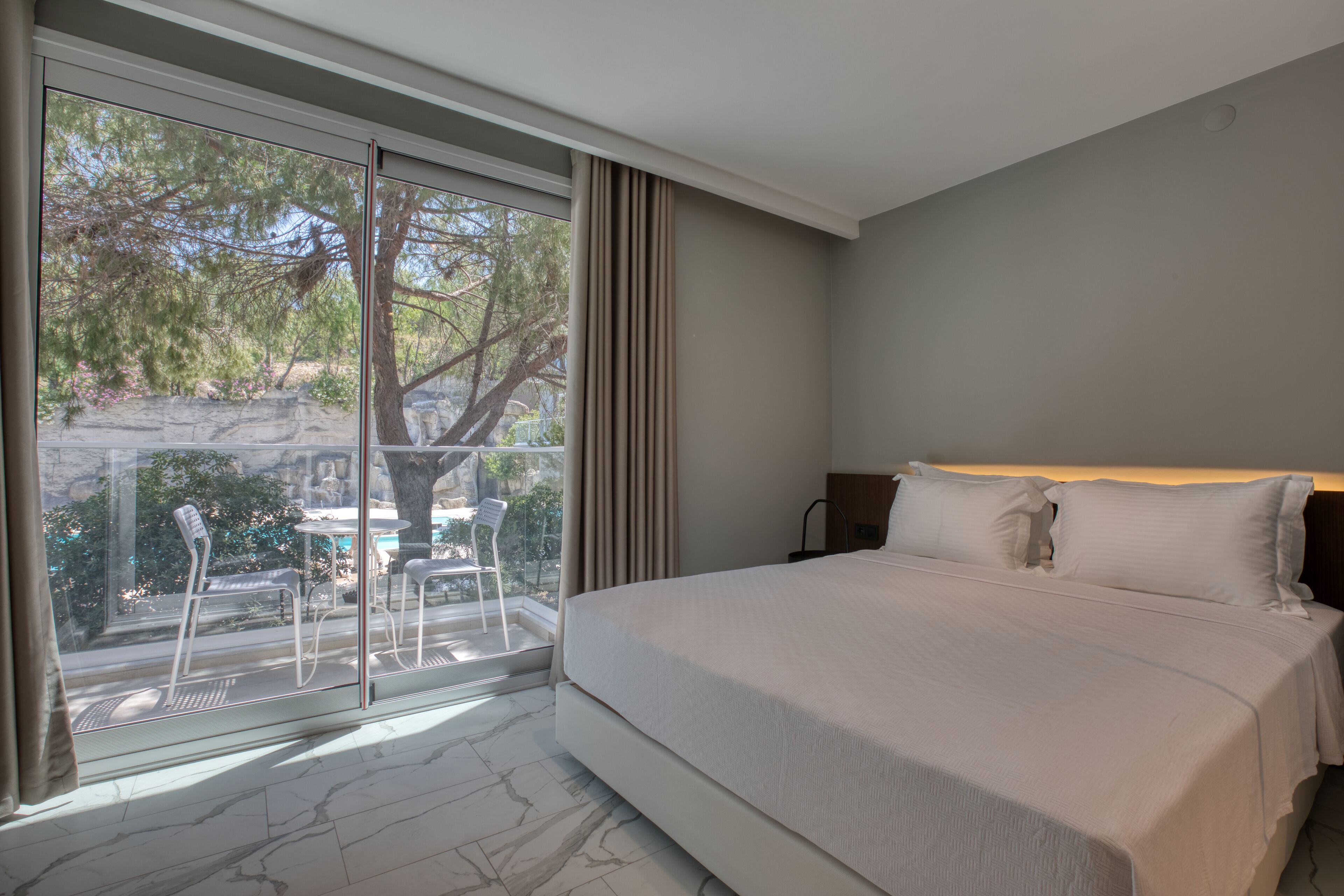 A hotel room with a large bed, a balcony with a table and chairs, and a view of trees and a building outside.