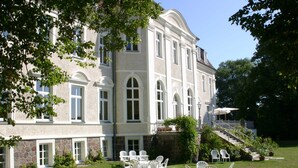 A large, historic building with a stone facade and multiple windows. There is a grassy lawn with outdoor furniture and a staircase leading to the upper levels. - The Gardener's House - Zinzow Castle (Boldekow)