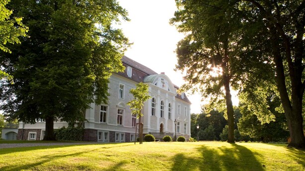 A large, historic building with a symmetrical facade, surrounded by a well-maintained lawn and mature trees. - The Gardener's House - Zinzow Castle (Boldekow)