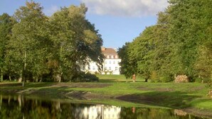 A large building with a symmetrical facade, surrounded by lush greenery and a calm body of water reflecting the structure. - The Gardener's House - Zinzow Castle (Boldekow)