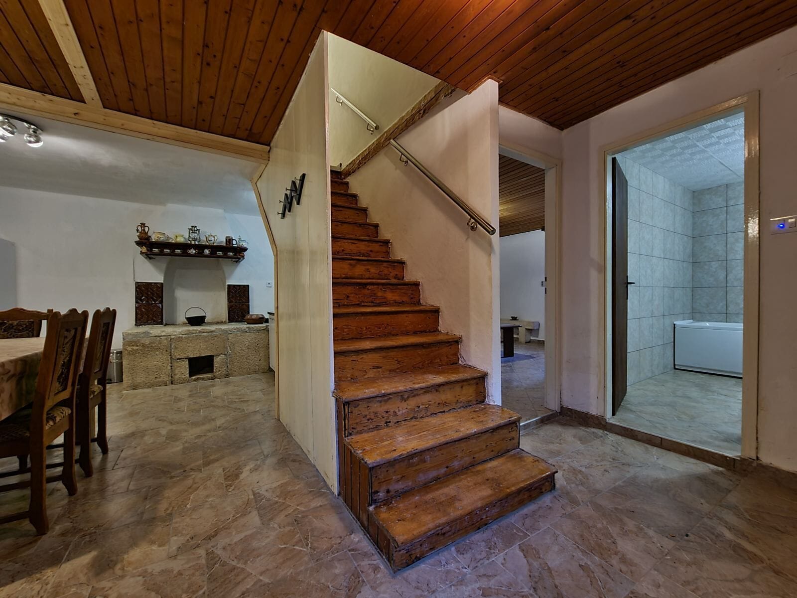 A wooden staircase leading to a second floor, a dining area with a stone table and chairs, and a bathroom with tiled walls and a bathtub.