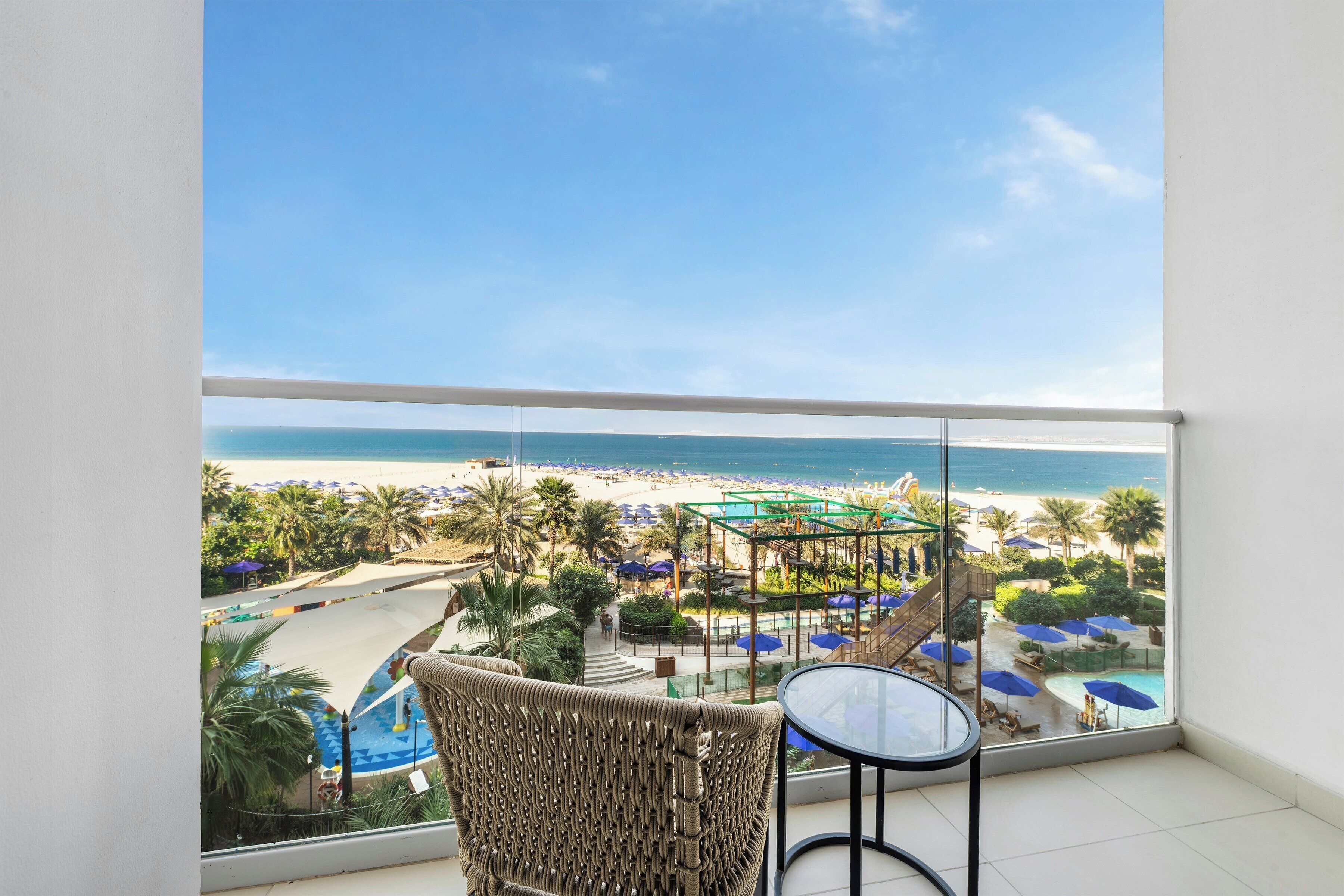 A balcony with a view of a beach, pool area, and lounge chairs.