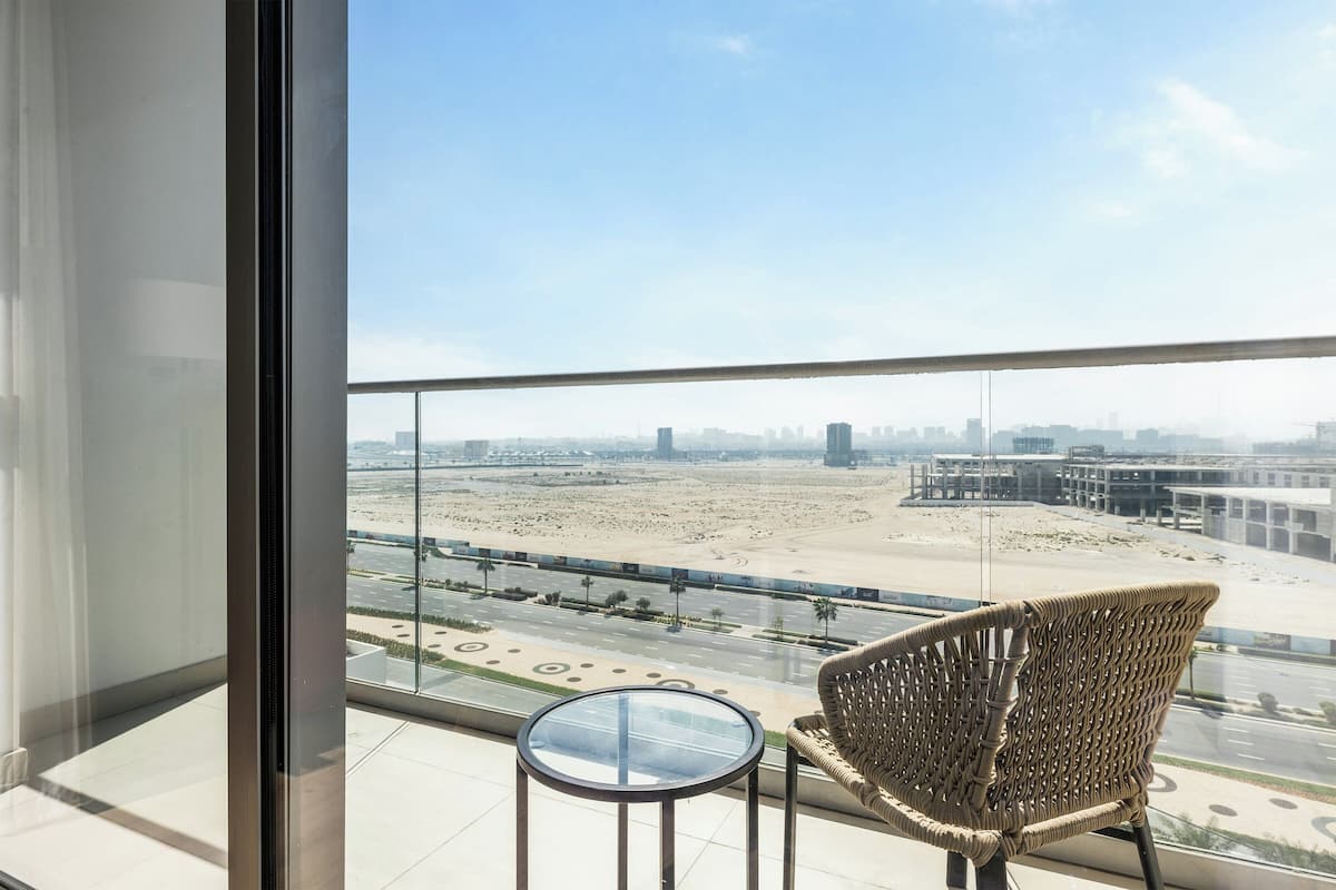 A balcony with a wicker chair and a small table, overlooking a cityscape and a desert area.