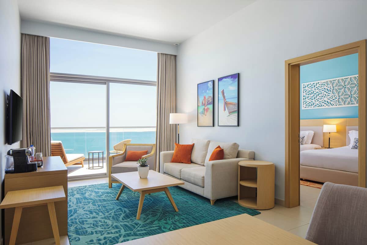 A hotel room with a large window, a sofa, a desk, a bed, and a TV.