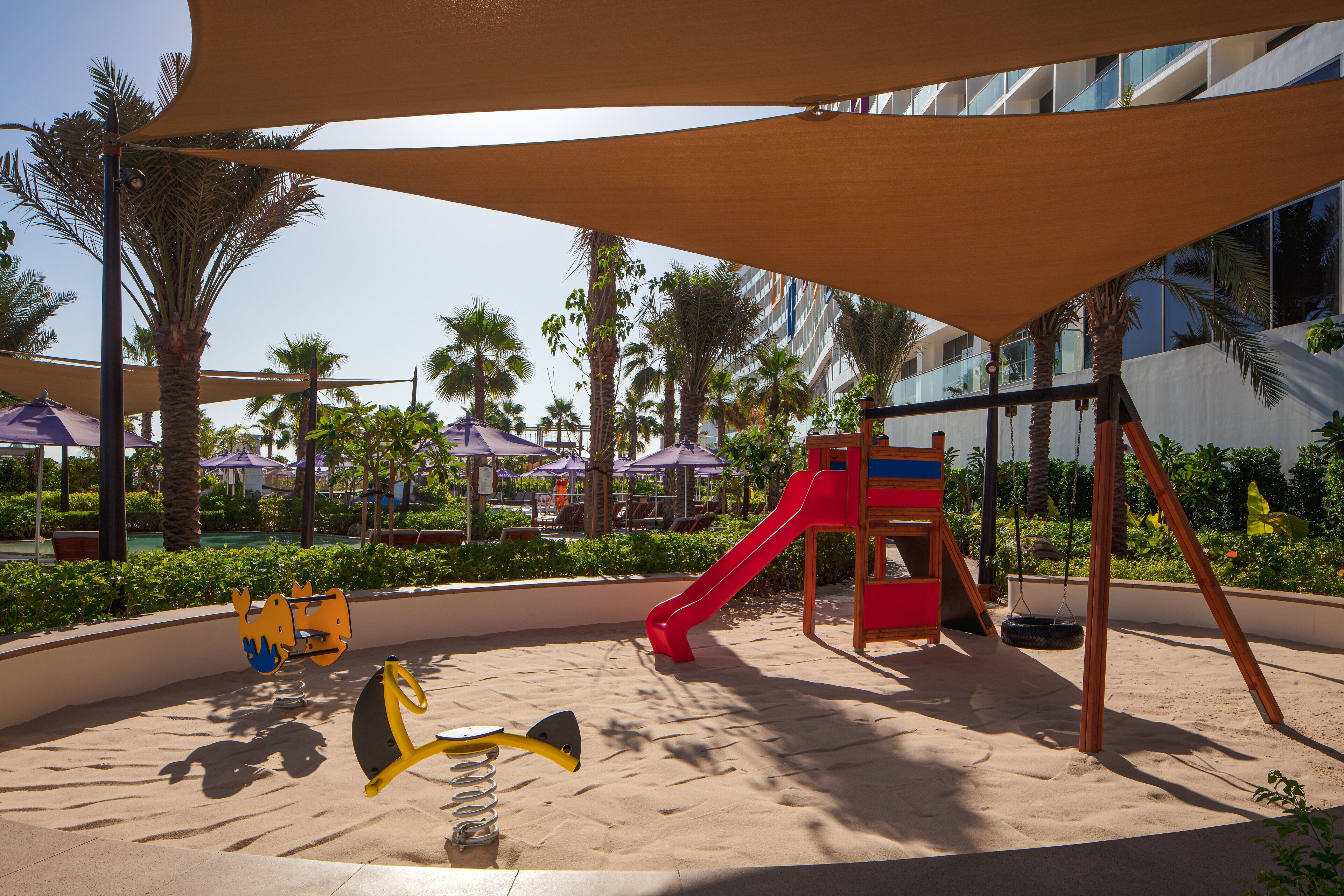 A playground with a slide and swings, surrounded by palm trees and a building in the background.