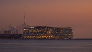 A modern hotel with a prominent glass facade, located near a waterfront with a city skyline in the background. - Centara Mirage Beach Resort Dubai (Dubai)